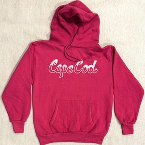 Könnte beinhalten: Ein roter Kapuzenpullover mit einem weißen "Cape Cod"-Aufdruck auf der Vorderseite. Der Pullover hat eine Kängurutasche und eine Kapuze mit Kordelzug.
