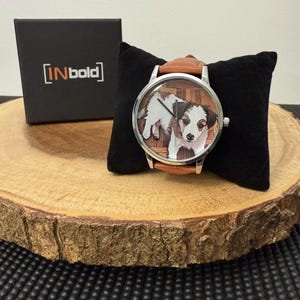 May include: A wristwatch with a brown leather band and a silver-colored case. The watch face features a color image of a puppy. A black box with the text "[INbold]" is visible in the background.