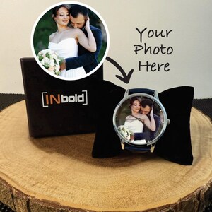 Puede incluir: Un reloj de pulsera personalizado con una esfera circular que muestra una fotograf&iacute;a impresa de una pareja con ropa de boda. El reloj tiene una caja de metal plateado y una correa azul oscuro, presentado sobre un coj&iacute;n de terciopelo negro.