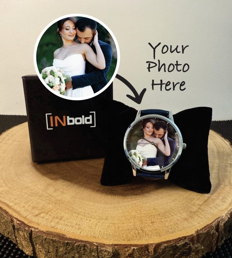 Puede incluir: Un reloj de pulsera personalizado con una esfera circular que muestra una fotograf&iacute;a impresa de una pareja con ropa de boda. El reloj tiene una caja de metal plateado y una correa azul oscuro, presentado sobre un coj&iacute;n de terciopelo negro.