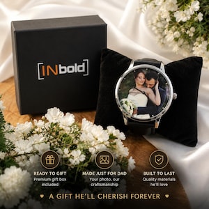 Puede incluir: Un reloj anal&oacute;gico de tono plateado con una fotograf&iacute;a personalizada de una pareja en la esfera, presentado sobre un coj&iacute;n de terciopelo negro junto a una caja de regalo negra con la etiqueta INbold. El reloj tiene una correa negra y esfera redonda.