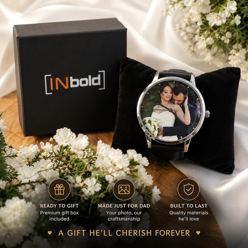 Puede incluir: Un reloj anal&oacute;gico de tono plateado con una fotograf&iacute;a personalizada de una pareja en la esfera, presentado sobre un coj&iacute;n de terciopelo negro junto a una caja de regalo negra con la etiqueta INbold. El reloj tiene una correa negra y esfera redonda.