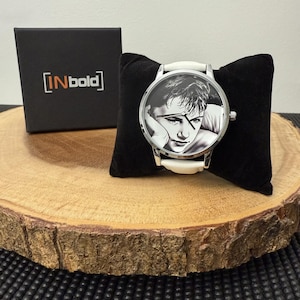 May include: A wristwatch with a black and white portrait on the face, a white band, and a silver case. The watch rests on a black velvet pillow, next to a black box with the text "[INbold]" in white. The watch is displayed on a wooden slice.