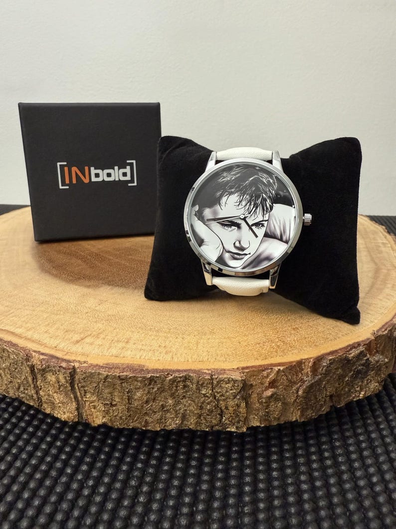 May include: A wristwatch with a black and white portrait on the face, a white band, and a silver case. The watch rests on a black velvet pillow, next to a black box with the text "[INbold]" in white. The watch is displayed on a wooden slice.