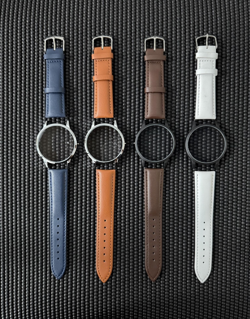 May include: Four watch faces with leather straps in blue, tan, brown, and white. The watch faces are silver and black. The straps have silver buckles and are arranged vertically against a textured black background.