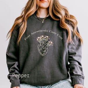 May include: A dark grey sweatshirt with the text "Post Anesthesia Care Unit" and a stylised heart with flowers. The text "Registered Nurse" is written on the heart. The word "Pepper" is written in white at the bottom of the image.