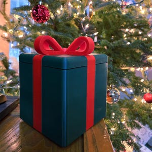 Puede incluir: Una caja de regalo verde oscuro con un lazo rojo en la parte superior y cintas rojas. La caja cuadrada está sobre una superficie de madera, con un árbol de Navidad al fondo.