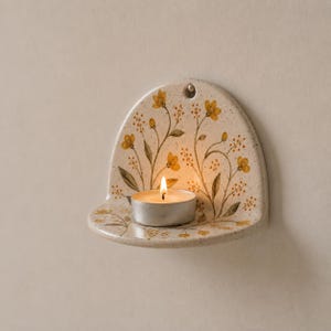 Puede incluir: Un portavelas de pared de cerámica en forma de semicírculo con un acabado crema moteado. La superficie presenta un diseño botánico pintado a mano de flores amarillas y tallos verdes. Una pequeña vela de té blanca encendida descansa en la repisa.