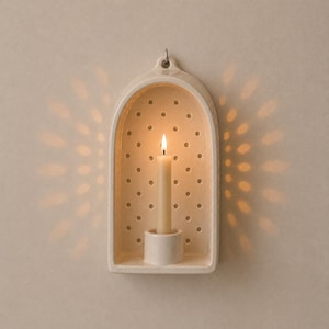 May include: An arched, off-white ceramic wall sconce featuring a perforated back panel with a grid of small circular holes. A single cream-colored taper candle sits in the central holder, casting a radiant, starburst-like light pattern.