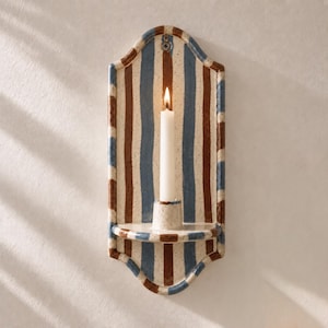May include: A wall-mounted candle sconce featuring a vertical striped pattern in shades of brown and blue on a speckled off-white background. The arched ceramic piece includes a small shelf with a central holder containing a single white taper candle.