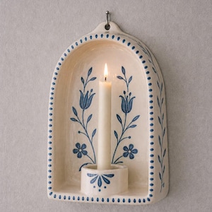 Può includere: Un portacandele da parete in ceramica color crema con una candela bianca accesa. Il portacandele presenta un motivo floreale blu e un bordo a pois. C'è un gancio di metallo nella parte superiore per appenderlo. Il portacandele è rotondo e decorato con motivi floreali blu.