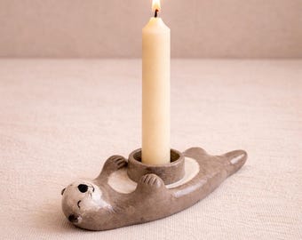 以下が含まれることがあります： グレーと白の色合いのセラミック製のカワウソ型キャンドルホルダー。クリーム色の火の灯ったキャンドルを保持しています。カワウソは仰向けに寝ており、前足を上げています。キャンドルは円形のホルダーの中央に配置されています。