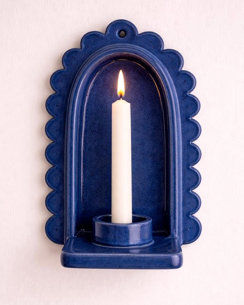 Peut inclure: Un photophore mural en c&eacute;ramique bleu marine avec un bord festonn&eacute;. Une bougie blanche est allum&eacute;e &agrave; l'int&eacute;rieur du photophore. Le photophore a un porte-bougie int&eacute;gr&eacute; et un petit trou en haut pour l'accrocher. Le design est simple et &eacute;l&eacute;gant.