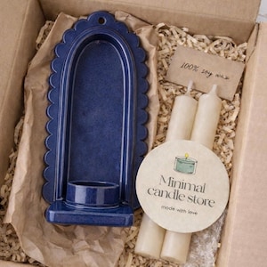 Peut inclure: Un bougeoir en c&eacute;ramique bleu marine avec un bord festonn&eacute;, emball&eacute; dans une bo&icirc;te en carton avec des bougies et une &eacute;tiquette indiquant "100% soy wax". Un autocollant rond indique "Minimal candle store made with love."