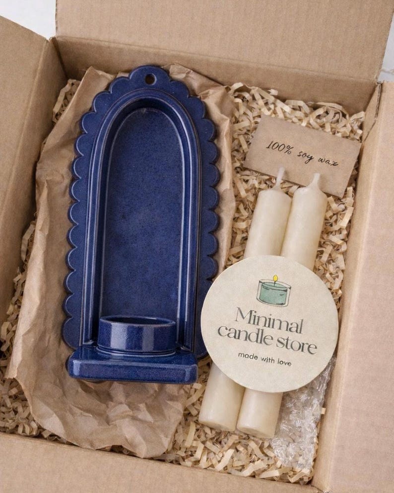 Peut inclure: Un bougeoir en c&eacute;ramique bleu marine avec un bord festonn&eacute;, emball&eacute; dans une bo&icirc;te en carton avec des bougies et une &eacute;tiquette indiquant "100% soy wax". Un autocollant rond indique "Minimal candle store made with love."