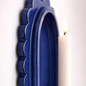 Peut inclure: Un bougeoir mural en c&eacute;ramique bleu avec un bord festonn&eacute; et un int&eacute;rieur en arc. Une bougie blanche est plac&eacute;e &agrave; l'int&eacute;rieur du bougeoir, avec une flamme allum&eacute;e. Le bougeoir est mont&eacute; sur un mur blanc.