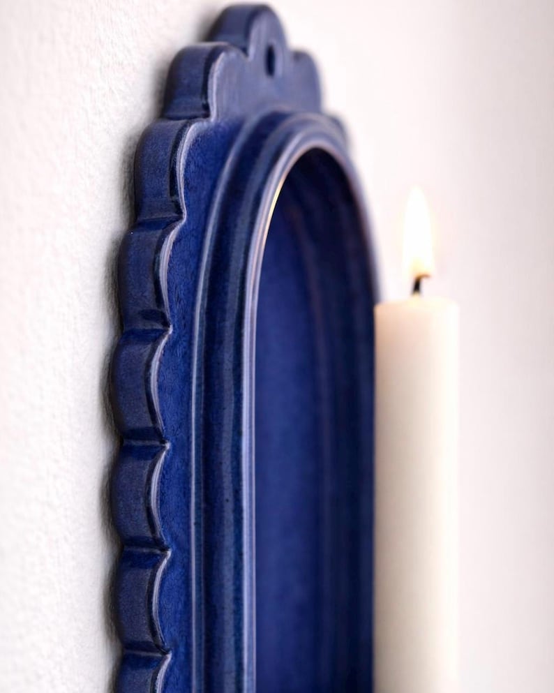 Peut inclure: Un bougeoir mural en c&eacute;ramique bleu avec un bord festonn&eacute; et un int&eacute;rieur en arc. Une bougie blanche est plac&eacute;e &agrave; l'int&eacute;rieur du bougeoir, avec une flamme allum&eacute;e. Le bougeoir est mont&eacute; sur un mur blanc.