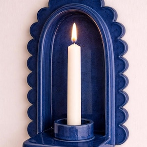 Peut inclure: Un bougeoir mural en c&eacute;ramique bleu avec un bord festonn&eacute;, contenant une bougie blanche allum&eacute;e. La bougie est plac&eacute;e dans un petit support rond. Le bougeoir a un design en arc courb&eacute;.