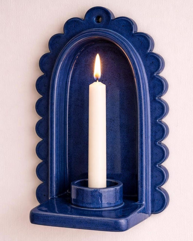 Peut inclure: Un bougeoir mural en c&eacute;ramique bleu avec un bord festonn&eacute;, contenant une bougie blanche allum&eacute;e. La bougie est plac&eacute;e dans un petit support rond. Le bougeoir a un design en arc courb&eacute;.