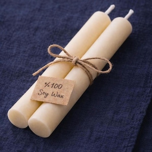 Peut inclure: Deux bougies cylindriques ivoire li&eacute;es avec de la ficelle. Une petite &eacute;tiquette rectangulaire avec le texte "%100 Soy Wax" est attach&eacute;e. Les bougies reposent sur un tissu bleu fonc&eacute;.
