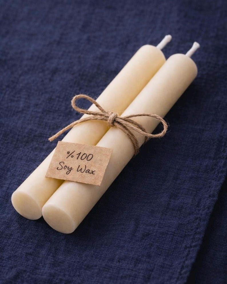 Peut inclure: Deux bougies cylindriques ivoire li&eacute;es avec de la ficelle. Une petite &eacute;tiquette rectangulaire avec le texte "%100 Soy Wax" est attach&eacute;e. Les bougies reposent sur un tissu bleu fonc&eacute;.