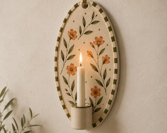 May include: An oval ceramic wall sconce featuring a cream-colored background with hand-painted orange flowers and green leaves. The border has a green and white checkered pattern. A cylindrical candle holder is attached at the bottom with a white candle.