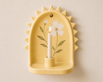 May include: A yellow ceramic wall sconce shaped like a sunburst arch featuring a painted white flower with green leaves. A small cylindrical candle holder is centered at the base, holding a single white taper candle with a lit flame.