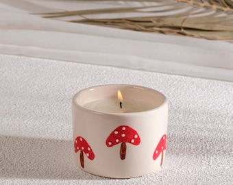 May include: A lit candle in a cream-colored ceramic container. The container is decorated with red mushroom illustrations with white polka dots and brown stems. The candle sits on a white surface.