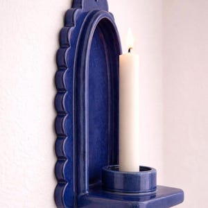 Peut inclure: Un bougeoir mural en c&eacute;ramique bleu cobalt avec un bord festonn&eacute;. Il contient une bougie blanche avec une flamme allum&eacute;e. Le bougeoir est une petite base ronde int&eacute;gr&eacute;e. Le bougeoir est fix&eacute; sur un mur blanc.
