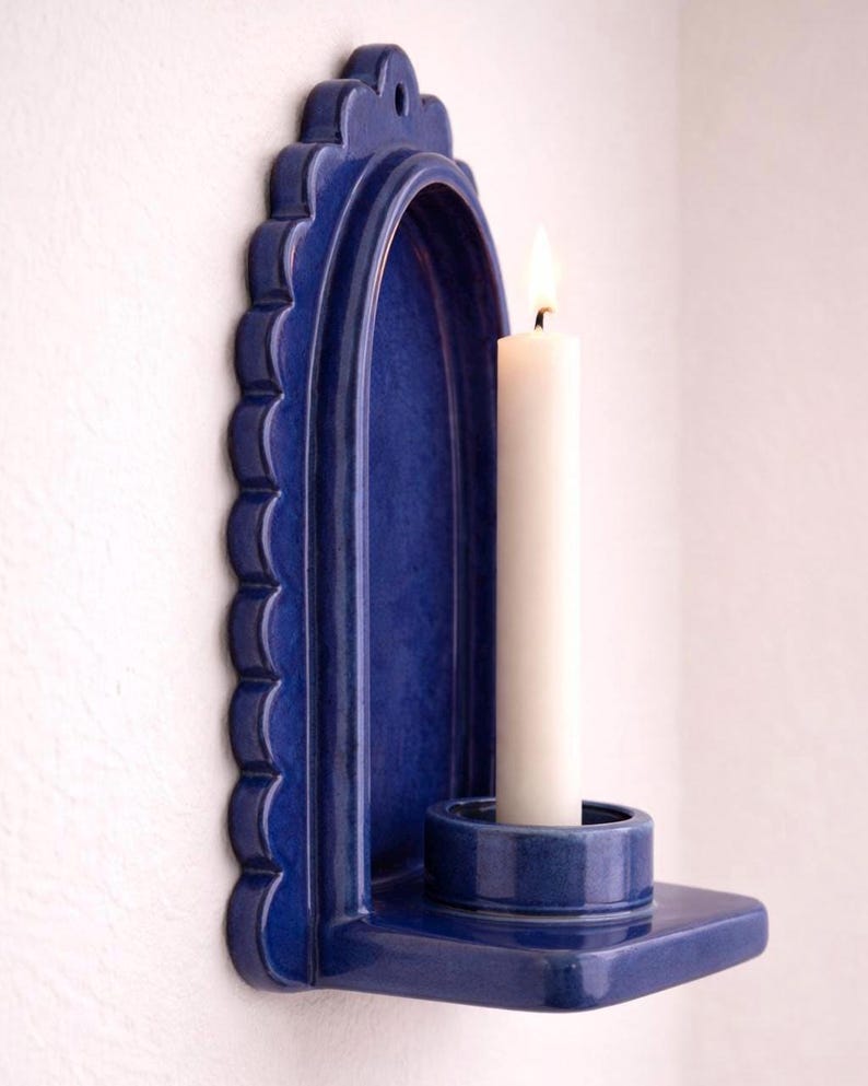 Peut inclure: Un bougeoir mural en c&eacute;ramique bleu cobalt avec un bord festonn&eacute;. Il contient une bougie blanche avec une flamme allum&eacute;e. Le bougeoir est une petite base ronde int&eacute;gr&eacute;e. Le bougeoir est fix&eacute; sur un mur blanc.