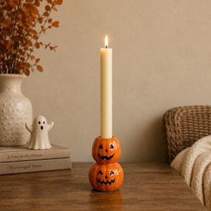 Puede incluir: Un portavelas de cerámica con forma de dos calabazas naranjas apiladas con caras de jack-o'-lantern pintadas en negro. Una vela cónica larga de color crema está insertada en la calabaza superior, con una llama encendida en la mecha.