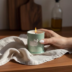 May include: A lit, pale green ceramic candle with a sheep design. The candle is in a cylindrical holder and sits on a white cloth. The flame is orange and yellow. The candle is on a wooden surface.