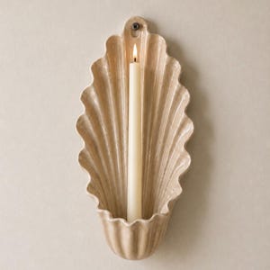 Op de afbeelding: Een beige schelpvormige wandkandelaar met een brandende, gebroken witte kaars. De kandelaar heeft een textuuroppervlak en een geschulpte rand. De kaars staat in het midden van de kandelaar en geeft een warme gloed. De kandelaar is aan een neutraal gekleurde muur bevestigd.