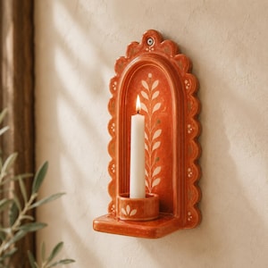 May include: A wall-mounted terracotta candle sconce with a scalloped edge and an arched backplate. The piece features a hand-painted white leaf vine pattern and a small circular candle holder at the base, holding a single white taper candle.