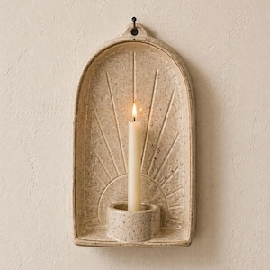 Peut inclure: Un bougeoir mural en céramique beige avec un motif de soleil. Le support a une petite base circulaire pour la bougie et un crochet pour l'accrocher. Une bougie allumée, de couleur blanc cassé, est dans le support.