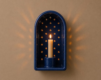 May include: A dark blue ceramic wall sconce with an arched top and a perforated back panel. The sconce features a central cylindrical holder containing a single lit cream-colored taper candle, casting light through the small circular holes.