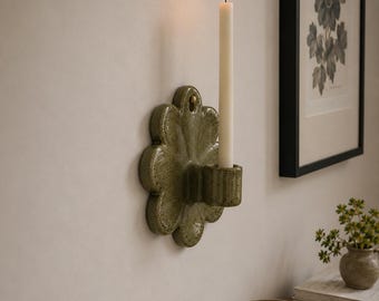 May include: A wall-mounted candle holder featuring a glossy, speckled olive green ceramic base shaped like a flower with rounded petals. A single, slender, off-white taper candle is inserted into the cylindrical holder, which is lit at the top.