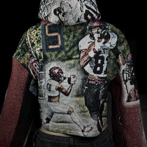 May include: Cropped hoodie featuring a tapestry design with football players in action. The hoodie has a hood and long sleeves with a burgundy-colored fabric. The main body of the hoodie displays a football game scene with players in white and red uniforms.