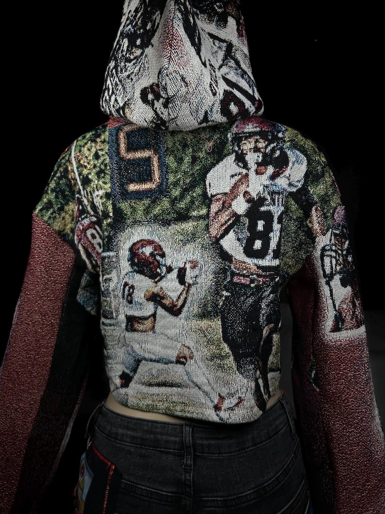 May include: Cropped hoodie featuring a tapestry design with football players in action. The hoodie has a hood and long sleeves with a burgundy-colored fabric. The main body of the hoodie displays a football game scene with players in white and red uniforms.