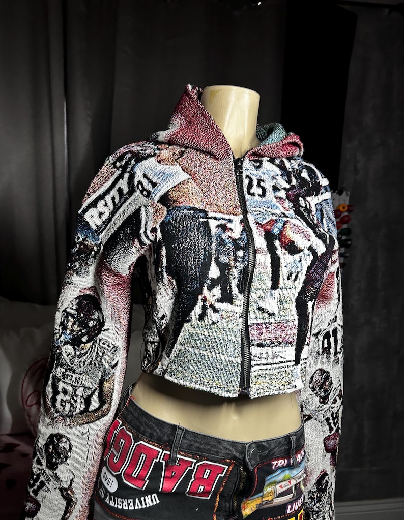 May include: Cropped tapestry jacket with a football theme. The jacket features a black zipper and a hood with a red and green lining. The fabric displays football players and the text "RSITY" and "25". The jacket is paired with denim shorts.