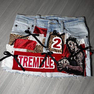 May include: Light blue denim skirt with patches of leopard print, red fabric, and a graphic of a person. The skirt has the word "TREMBLE" in white on a red patch and the number "2". Black ribbons are tied on the skirt.