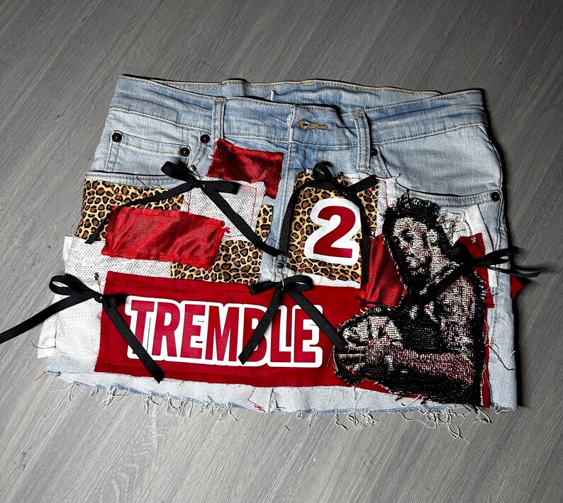 May include: Light blue denim skirt with patches of leopard print, red fabric, and a graphic of a person. The skirt has the word "TREMBLE" in white on a red patch and the number "2". Black ribbons are tied on the skirt.