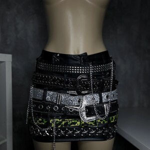 May include: Black mini skirt with multiple layered belts. The belts feature silver studs, rhinestones, and chain details. One belt has a green and black camouflage pattern. The skirt is displayed on a mannequin.