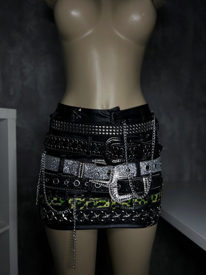 May include: Black mini skirt with multiple layered belts. The belts feature silver studs, rhinestones, and chain details. One belt has a green and black camouflage pattern. The skirt is displayed on a mannequin.
