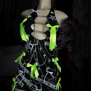 May include: Black halter dress with silver chain detailing, cut-outs, and neon green bows. The mini dress is adorned with silver star and cross embellishments. The dress is displayed on a mannequin.