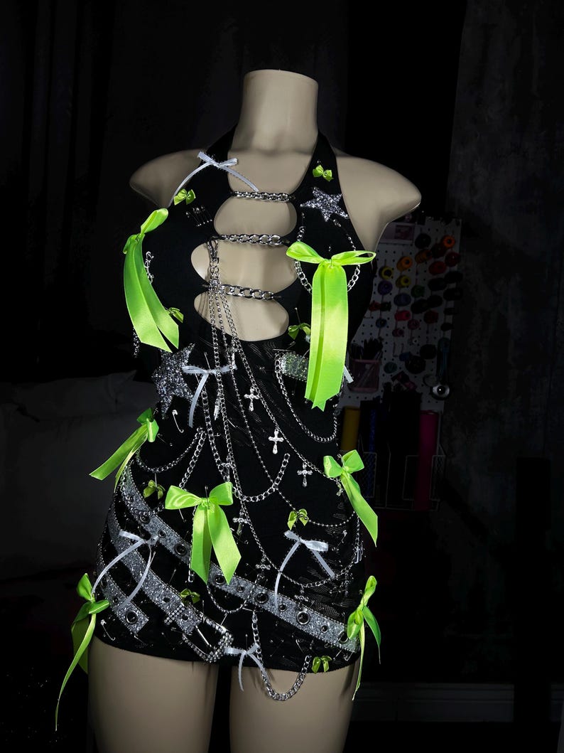 May include: Black halter dress with silver chain detailing, cut-outs, and neon green bows. The mini dress is adorned with silver star and cross embellishments. The dress is displayed on a mannequin.