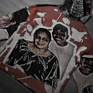 May include: A woven textile with a portrait of two smiling people. The artwork is in shades of red, white, and black, with a textured appearance. The textile appears to be a hoodie or jacket.