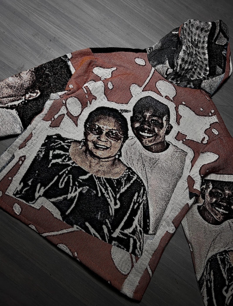 May include: A woven textile with a portrait of two smiling people. The artwork is in shades of red, white, and black, with a textured appearance. The textile appears to be a hoodie or jacket.