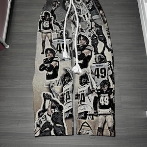 May include: Tapestry-style sweatpants featuring a repeating pattern of football players in action. The pants are primarily beige and brown, with a white drawstring. The players are wearing black and white uniforms with the number 49.