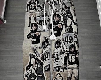 May include: Woven pants featuring a collage of football players in black and white uniforms with the number 49 and the word Purdue printed on them. The pants have a tan and brown gradient background, a thick white drawstring at the waist, and a straight-leg fit.